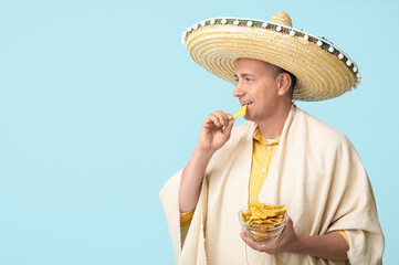 Wall Mural - Handsome mature Mexican man in sombrero hat with bowl of tasty nachos on blue background