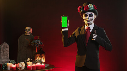 Wall Mural - Horror lady of dead showing smartphone with greenscreen in studio, holding isolated chroma key display background. Scary model using blank mockup copyspace template, wearing suit.