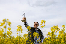 Adult Man Triumphantly Holds Wind Turbine Model In Yellow Raps Field.