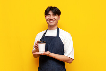 Wall Mural - young asian guy barista in apron holding glass of coffee on yellow isolated background, korean male waiter in uniform