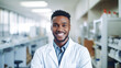 Portrait of black young man wearing lab coat and smiling at camera in workshop of pharmaceutical factory.

