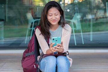 Beautiful Chinese teenage student smiling using social media app on mobile phone outdoors. Girl chatting on mobile phone