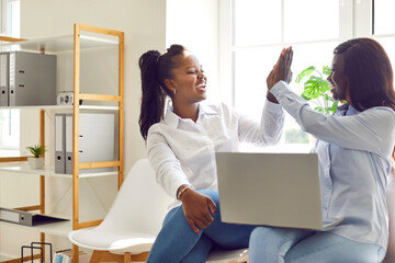 Two happy young smiling African American business women coworkers entrepreneurs sitting on chairs with laptop computer in office workplace, smiling and giving each other high five. Teamwork concept