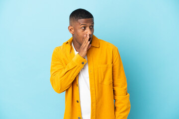 Young latin man isolated on blue background whispering something with surprise gesture while looking to the side