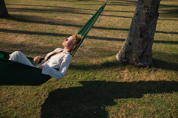 A young girl with curly blond hair lies in a hammock in nature among palm trees and enjoys life.