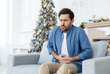A Young Man Suffers From Severe Abdominal Pain During The Christmas Holidays. He Sits At Home On The Sofa Near The Christmas Tree And Holds His Body With His Hands, Grimacing From Spasms