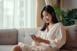 © JAYDESIGNZ - Happy young asian woman sitting on sofa holding mobile phone using cellphone technology doing ecommerce shopping, buying online, texting messages relaxing on couch in cozy living room at home