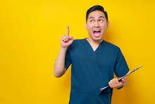 Excited Professional Young Asian Male Doctor Or Nurse Wearing A Blue Uniform Holding Clipboard And Pointing Finger Up, Having Great Ideas Isolated On Yellow Background. Healthcare Medicine Concept