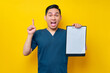 Excited professional young Asian male doctor or nurse wearing a blue uniform holding clipboard with blank paper and pointing finger up, having good ideas isolated on yellow background