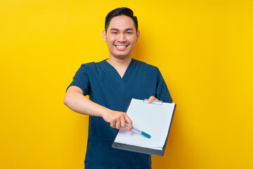 Wall Mural - Professional young Asian male doctor or nurse wearing a blue uniform pointing with pen at blank paper on clipboard for patient signature approval isolated on yellow background