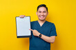© Bangun Stock Photo - Smiling professional young Asian male doctor or nurse wearing a blue uniform pointing finger at clipboard with blank paper isolated on yellow background. Healthcare medicine concept