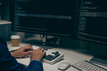 Male programmer working on desktop computer at white table in office, coding at night on simulated laptop screen for network, malware or office software.