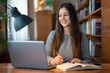 © Vitalii - Cute young student girl sitting at a table with laptop in front of her, writing something in the notebook. Concept of education, studying in a library, modern students