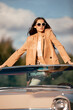 © Lui Shtein - A beautiful woman sits in a convertible in a field. Dressed in a beige jacket and white trousers and black sunglasses. Beautiful and stylish look
