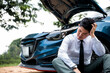 © sorapop - Man sitting near his broken car with raised hood on rural road, calling for roadside assistance. Concept of car trouble and frustration.