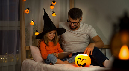 Father and little daughter in witch black hat watch horror film on tablet in dark with orange pumpkin lamp. Halloween celebration