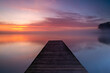 A wooden pier extends into the lake, where a layer of fog forms above the calm water. The rising sun beautifully colors the sky.