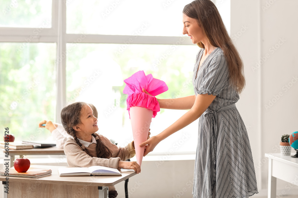 Teacher greeting her happy pupil with pink school cone in classroom