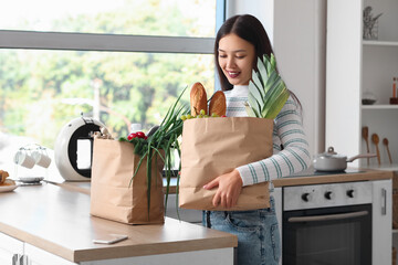 Wall Mural - Young Asian woman with mobile phone and shopping bags full of fresh food at table in kitchen