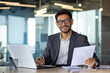 © Liubomir - Portrait of young successful Indian businessman financier, man smiling and looking at camera, working sitting at laptop, holding reports, papers and contracts, satisfied investor entrepreneur.