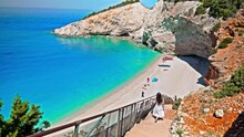 A Beautiful Woman In A White Dress Walks Down The Stairs To See Porto Katsiki Beach. Female Tourist Relax At The Narrow Beach With Picturesque Views, Turquoise Water Closely Bordered By Steep Cliffs.