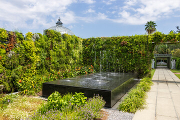 Wall Mural - A black marble water fountain surrounded by lush green trees and plants and colorful flowers with blue sky and clouds at New Orleans Botanical Garden in New Orleans Louisiana USA