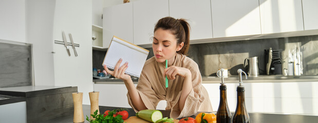 Wall Mural - Portrait of woman checking grocery list, looking at vegetables, holding notebook, reading recipe while cooking meal in the kitchen, chopping tomatoes and zucchini