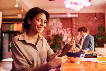 Poster - Cheerful woman with mobile phone communicating in video chat while sitting in Japanese cafe or restaurant at lunch break and eating ramen