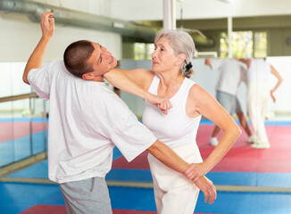 Wall Mural - Focused elderly woman performing elbow strike and wristlock, painful control move to immobilize male opponent during self defense training in gym