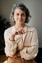 Poster - Portrait of experienced female chief executive officer with eyeglasses in hand looking at camera while standing against grey wall of office