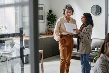 Poster - Young Hispanic businesswoman with papers making report of financial data to experienced female chief executive officer of company
