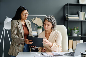 Poster - Confident mature female boss looking through document held by young secretary and checking points while sitting by her workplace