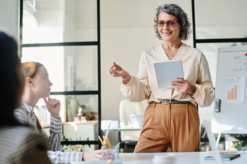 Poster - Happy confident mature businesswoman with tablet explaining something to young subordinates sitting in front of her during training or seminar