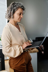 Poster - Serious mature female analyst in quiet luxury apparel looking at screen of laptop and working in the net or organizing work while standing in office
