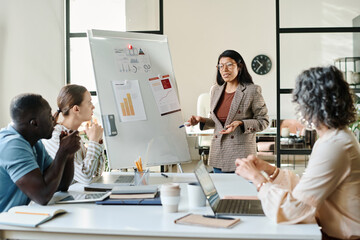 Young confident economist standing by whiteboard in front of colleagues during explanation of financial charts and diagrams on papers