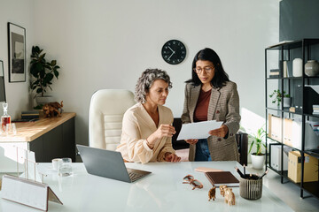 Wall Mural - Mature female boss looking through documents held by young Hispanic secretary standing next to her and consulting about some points