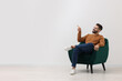 © New Africa - Handsome man pointing at something while sitting in armchair near white wall indoors, space for text