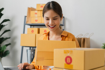 Wall Mural - Startup small business, Young Asian woman holding packing boxes and checking online order on digital laptop for products to send to customers. working at the home office.