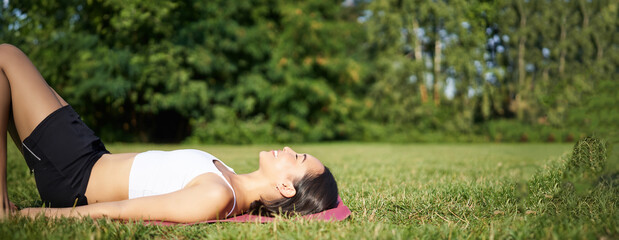 Wall Mural - Young fitness girl lying on sport mat on lawn, breathing and meditating in park in sportswear