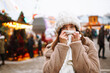 Young woman in winter style clothes posing at festive street market. Woman enjoying winter moments. Beautiful light decorations. Festive Christmas fair, winter holidays concept. 