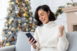 Happy young woman sitting at home on sofa near Christmas tree and using mobile phone, happy with success and good news for New Year holidays, showing victory gesture with hand
