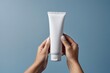 © Mykola - Female hand holding a white tube of cosmetic cream on a blue background, generative ai