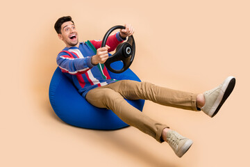 Poster - Full body length photo of young guy sit bean bag and drive steering wheel automobile simulation service isolated on beige color background