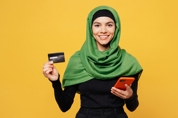 Wall Mural - Young muslim woman wearing green hijab abaya black clothes use mobile cell phone credit bank card shopping online isolated on plain yellow background People uae middle eastern islam religious concept