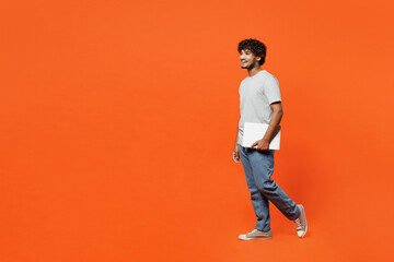 Wall Mural - Full body side view young happy IT Indian man he wearing t-shirt casual clothes hold closed laptop pc computer walking going isolated on orange red color background studio portrait. Lifestyle concept.
