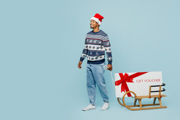 Sticker - Full body young man wear sweater Santa hat posing carry on sled store gift certificate coupon voucher card isolated on plain blue background. Happy New Year 2024 celebration Christmas holiday concept.