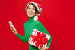 © ViDi Studio - Full body young woman wears green turtleneck Santa hat posing hold mobile cell phone present box with gift ribbon bow look aside isolated on plain red background Happy New Year 2024 Christmas concept
