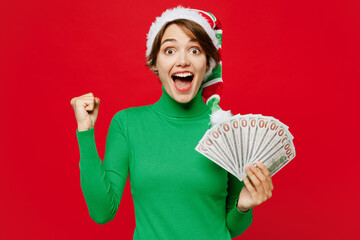 Wall Mural - Young woman wear green turtleneck Santa hat posing hold in hand fan of cash money in dollar banknotes do winner gesture isolated on plain red background. Happy New Year 2024 Christmas holiday concept.