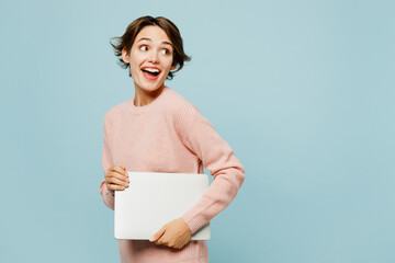 Side view young IT woman she wear beige knitted sweater casual clothes hold closed laptop pc computer look aside isolated on plain pastel light blue cyan background studio portrait. Lifestyle concept