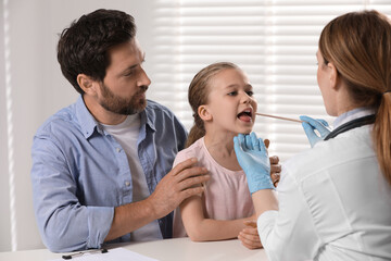 Canvas Print - Doctor examining girl`s oral cavity with tongue depressor near her father indoors